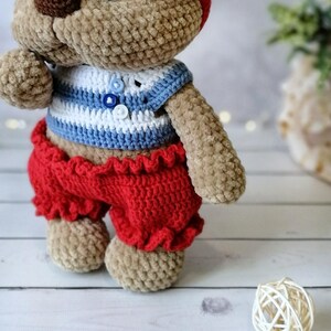 May include: A crocheted teddy bear wearing a blue and white striped shirt and red pants with a ruffled hem.