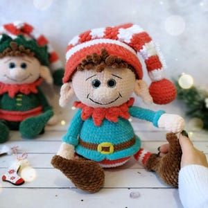May include: A handmade crochet elf doll with a red and white striped hat, a turquoise shirt, and brown shoes. The elf has a smiling face and is wearing a red collar. The doll is a festive holiday decoration.