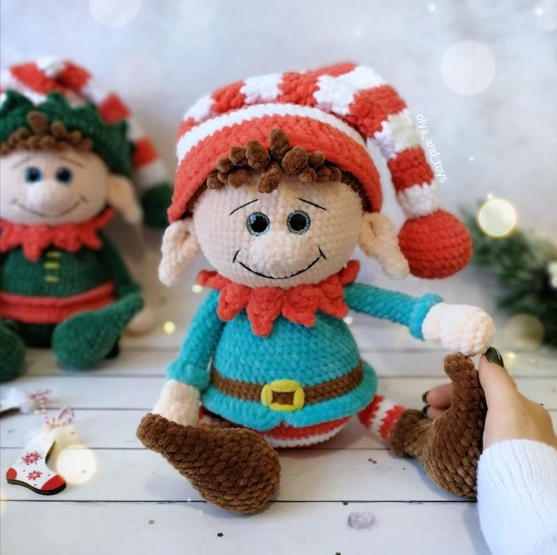 May include: A handmade crochet elf doll with a red and white striped hat, a turquoise shirt, and brown shoes. The elf has a smiling face and is wearing a red collar. The doll is a festive holiday decoration.