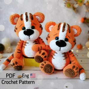 May include: Two crocheted tiger plushies, one orange and white with brown stripes, the other orange and white with brown stripes, sitting on a wooden surface. The plushies have black eyes and blue eyes, and both have brown noses. The plushies are sitting on a yellow surface. The text "PDF Eng Crochet Pattern" is at the bottom of the image.