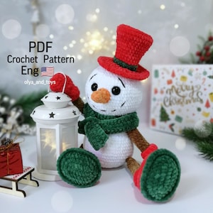 Op de afbeelding: Een gehaakte sneeuwpop met een rode hoge hoed, groene sjaal en rode wanten. De sneeuwpop heeft een wortelneus en zwarte ogen. Hij houdt een witte lantaarn vast. De tekst "PDF Crochet Pattern Eng" is zichtbaar.