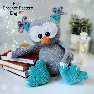 May include: A crocheted gray owl with teal wings and feet. The owl has white eyes and an orange beak. The owl is sitting on a stack of books with the text "olya_and_toys" visible.
