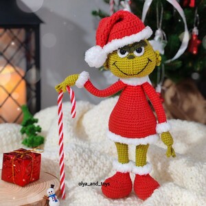 Puede incluir: Mu&ntilde;eco Grinch tejido a ganchillo con gorro de Pap&aacute; Noel rojo y atuendo a juego con ribetes blancos, sosteniendo un bast&oacute;n de caramelo. El mu&ntilde;eco tiene una cara amarilla y ojos verdes. Un peque&ntilde;o mu&ntilde;eco de nieve y un regalo envuelto est&aacute;n en primer plano.