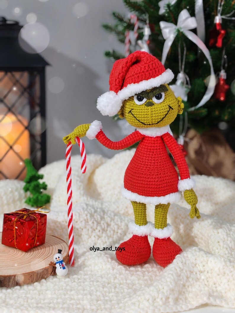 Puede incluir: Mu&ntilde;eco Grinch tejido a ganchillo con gorro de Pap&aacute; Noel rojo y atuendo a juego con ribetes blancos, sosteniendo un bast&oacute;n de caramelo. El mu&ntilde;eco tiene una cara amarilla y ojos verdes. Un peque&ntilde;o mu&ntilde;eco de nieve y un regalo envuelto est&aacute;n en primer plano.