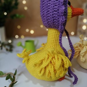 May include: A crocheted yellow duck wearing a purple crocheted bonnet. The duck has a long, thin beak and a fluffy tail.