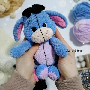 May include: A blue crocheted stuffed animal resembling a donkey with pink ears, black eyes, and a black bow on its head. The animal is being held by a person's hands.