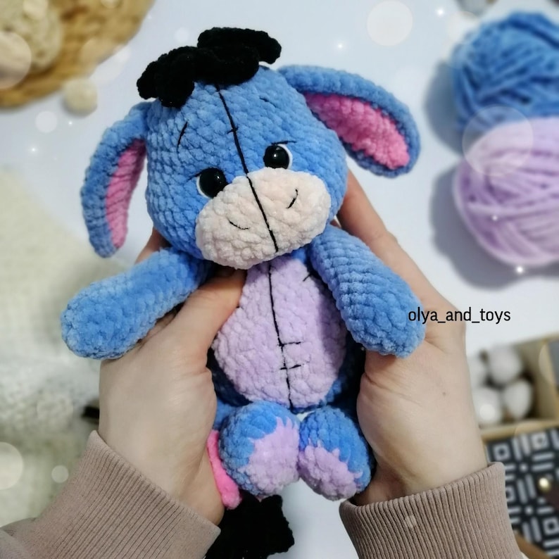 May include: A blue crocheted stuffed animal resembling a donkey with pink ears, black eyes, and a black bow on its head. The animal is being held by a person's hands.