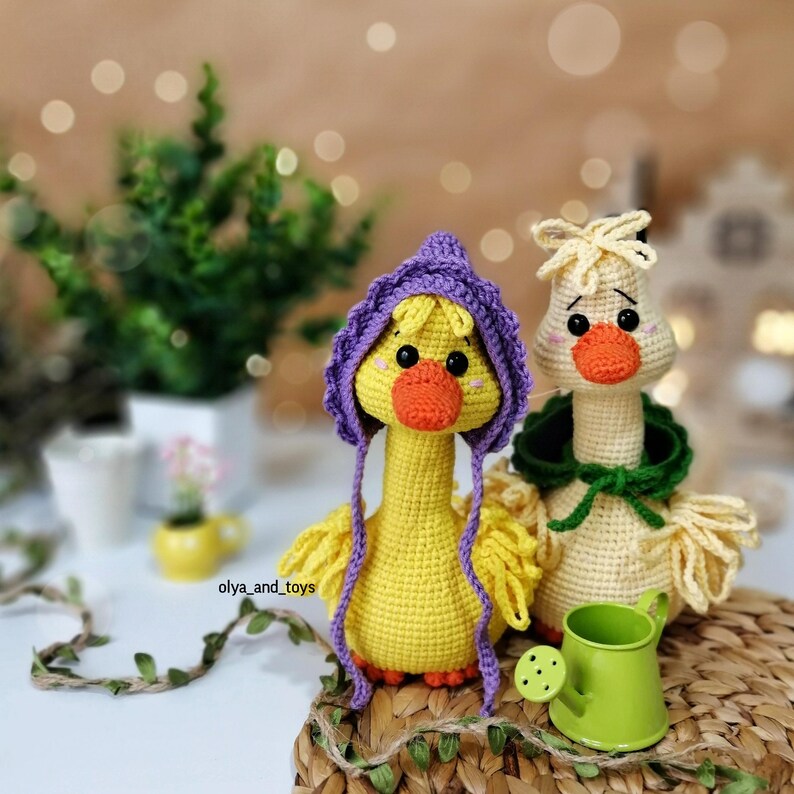 May include: Two crocheted yellow goose plushies with long necks. One goose is wearing a purple hooded hat and the other is wearing a green hooded cape. The geese are sitting on a woven basket with a green watering can in front of them.