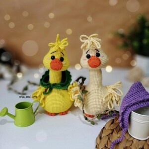 May include: Two crocheted yellow and cream colored goose plushies with orange beaks and black eyes. The goose on the left has a green crocheted scarf around its neck. The goose on the right has a purple crocheted hat.