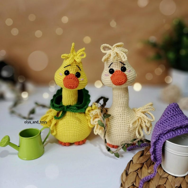 May include: Two crocheted yellow and cream colored goose plushies with orange beaks and black eyes. The goose on the left has a green crocheted scarf around its neck. The goose on the right has a purple crocheted hat.
