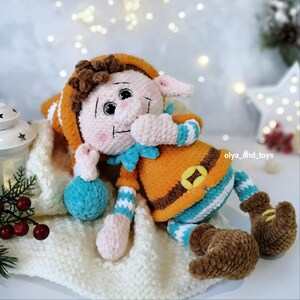 May include: A handmade crochet elf doll with a smiling face, wearing an orange outfit with a blue collar and striped sleeves and legs. The doll has brown hair and brown boots. It is lying on a white knitted blanket.