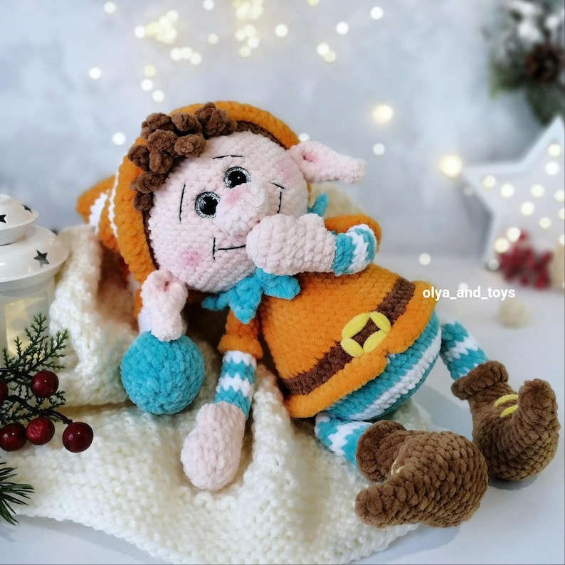 May include: A handmade crochet elf doll with a smiling face, wearing an orange outfit with a blue collar and striped sleeves and legs. The doll has brown hair and brown boots. It is lying on a white knitted blanket.
