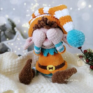 May include: A handmade crochet elf doll. It features an orange and white striped hat, orange tunic, and brown shoes. The doll has a blue pom-pom on the hat and blue and white striped cuffs. The doll's face is pink and the eyes are covered.