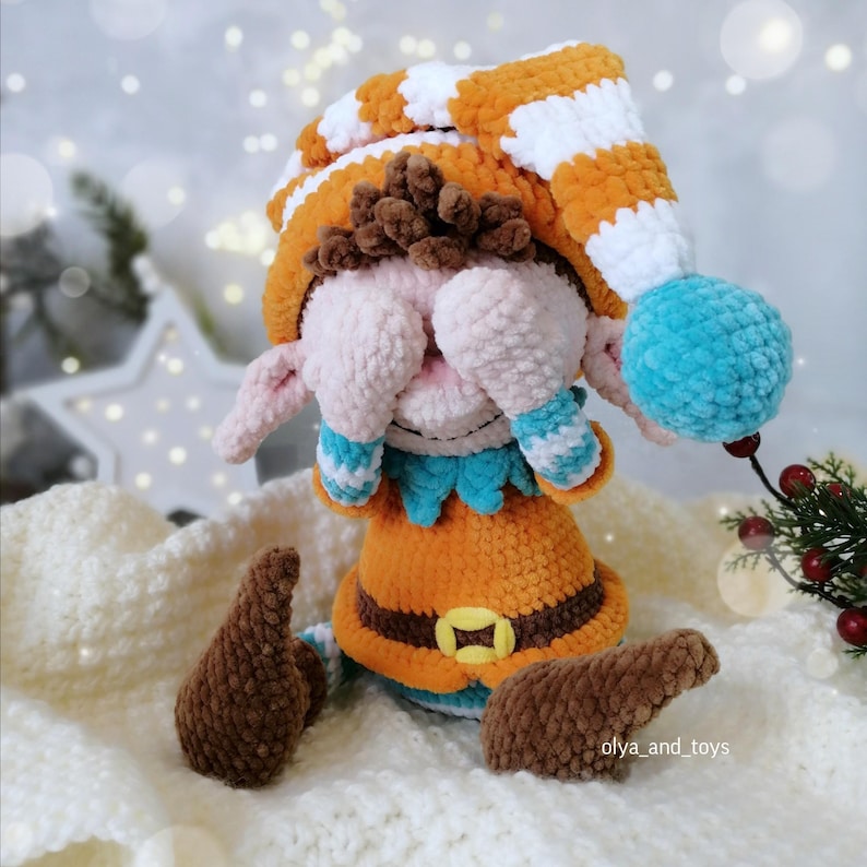 May include: A handmade crochet elf doll. It features an orange and white striped hat, orange tunic, and brown shoes. The doll has a blue pom-pom on the hat and blue and white striped cuffs. The doll's face is pink and the eyes are covered.