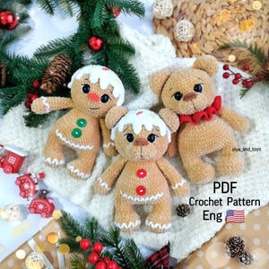 Puede incluir: Tres juguetes de ganchillo hechos a mano: dos hombres de jengibre y un oso de peluche. Los hombres de jengibre tienen glaseado blanco, detalles rojos y verdes. El oso de peluche tiene un cuello con volantes rojos. Los juguetes están sobre una superficie de punto blanca, con decoraciones navideñas.