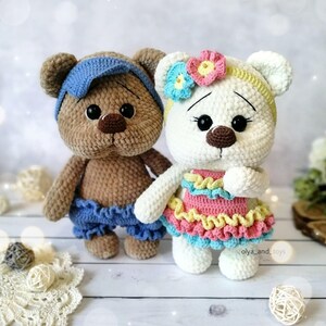 May include: Two crocheted teddy bears, one brown with a blue headband and blue shorts, the other white with a yellow headband and a pink and blue dress.