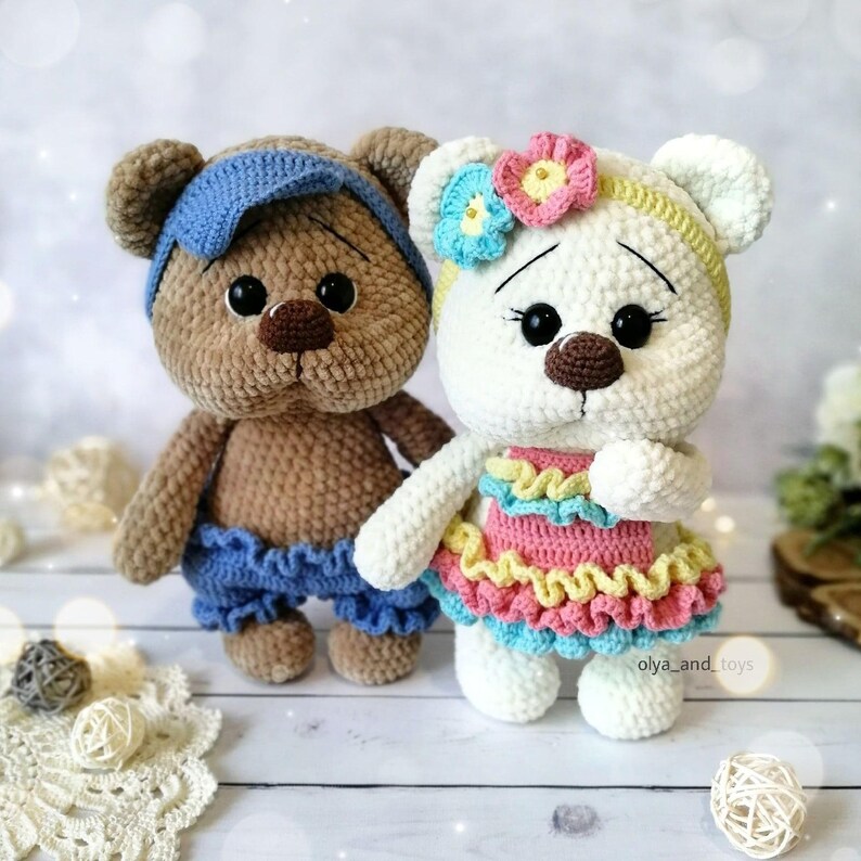 May include: Two crocheted teddy bears, one brown with a blue headband and blue shorts, the other white with a yellow headband and a pink and blue dress.
