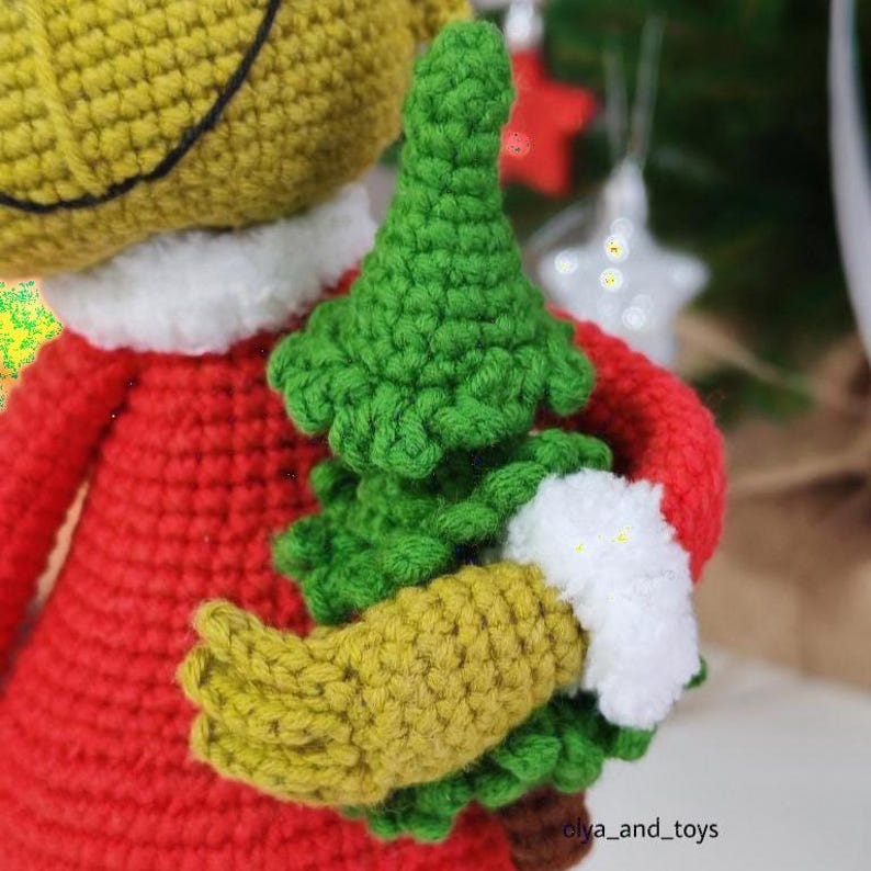 Puede incluir: Mu&ntilde;eca de ganchillo hecha a mano con un atuendo rojo, sosteniendo un &aacute;rbol de Navidad verde. La mu&ntilde;eca tiene una cara amarilla y un cuello blanco. La mano de la mu&ntilde;eca es amarilla y el &aacute;rbol es verde. Decoraci&oacute;n festiva.