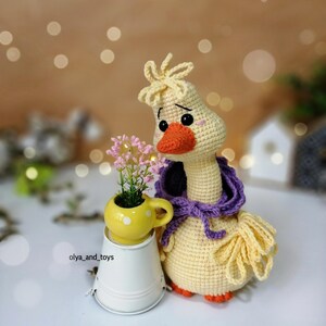 May include: A crocheted yellow goose toy with a purple scarf, standing next to a yellow ceramic pot with pink flowers. The goose has black eyes and an orange beak.