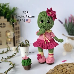 May include: Crochet pattern for a green cactus doll wearing a pink dress and a flower on its head. The doll is standing on a woven surface. The pattern is available in English. PDF Crochet Pattern Eng.
