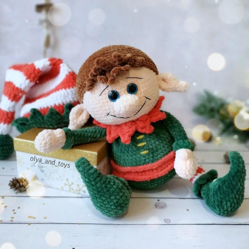 May include: A handmade crochet elf doll in green and red, with brown hair and blue eyes. The elf sits beside a gift box and a striped hat. The doll has a smiling face, a festive holiday decoration. The doll is a charming Christmas ornament.