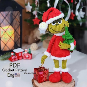 Puede incluir: Mu&ntilde;eco Grinch tejido a ganchillo con un traje de Pap&aacute; Noel rojo con ribetes blancos y un gorro a juego. El Grinch sostiene un peque&ntilde;o &aacute;rbol de Navidad verde y est&aacute; cerca de una caja de regalo roja. El texto "PDF Crochet Pattern Eng" es visible.