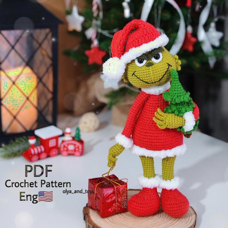 Puede incluir: Mu&ntilde;eco Grinch tejido a ganchillo con un traje de Pap&aacute; Noel rojo con ribetes blancos y un gorro a juego. El Grinch sostiene un peque&ntilde;o &aacute;rbol de Navidad verde y est&aacute; cerca de una caja de regalo roja. El texto "PDF Crochet Pattern Eng" es visible.