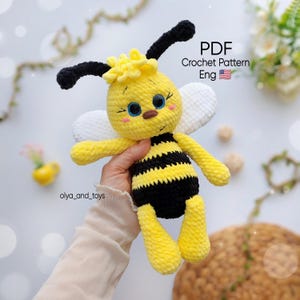 Pode incluir: Um brinquedo de abelha em crochê com corpo listrado de amarelo e preto, asas brancas e antenas pretas. A cabeça apresenta olhos azuis, nariz marrom e bochechas rosadas. O texto na imagem diz PDF Crochet Pattern Eng com uma bandeira dos EUA.