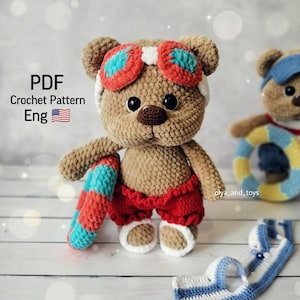 May include: Crocheted teddy bear wearing red shorts, blue and red goggles, and holding a blue and red life preserver.  The bear is standing on a white wooden surface.  PDF Crochet Pattern Eng.