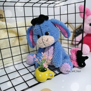 May include: A blue and purple crocheted stuffed animal resembling a donkey with a black bow on its head. The animal is sitting in a wire basket with a small yellow cup with white polka dots and pink flowers.