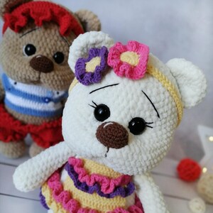 May include: Two crocheted teddy bears wearing colorful outfits. The white bear has a yellow headband with pink and purple flowers. The brown bear is wearing a blue and white striped shirt and a red skirt.