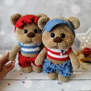 May include: Two crocheted teddy bears wearing sailor-themed outfits. The bear on the left is wearing a red and white striped shirt, red shorts, and a red headband. The bear on the right is wearing a red and white striped shirt, blue shorts, and a blue headband.