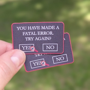 May include: Two rectangular stickers with a black background and red border. The text reads: YOU HAVE MADE A FATAL ERROR. TRY AGAIN? with two white boxes labelled YES and NO. A small red magnifying glass icon is positioned over the YES box.
