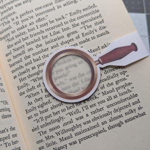 May include: A magnifying glass bookmark with a brown handle and a gold rim. The magnifying glass is over a page of a book with the text "broken in, are you going to jewels?" visible through the glass.