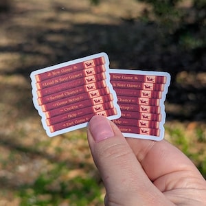 May include: Two stickers depicting a stack of books with game menu options such as "New Game" and "Load & Save Game." The stickers have a burgundy and gold colour scheme, with white text. The stickers are held in a person's hand.