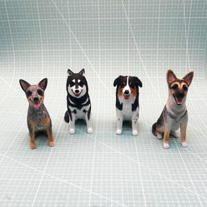 Könnte beinhalten: Vier 3D-gedruckte Figuren von Hunden in sitzender Pose. Die Hunde sind ein schwarz-weißer Husky, ein Blue Merle Australian Shepherd, ein Deutscher Schäferhund und ein Blue Heeler.