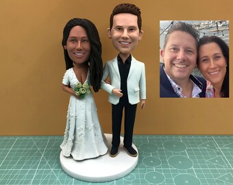 May include: A wedding cake topper featuring a bride and groom figurine. The bride wears a white dress and holds a bouquet. The groom is in a white jacket and black pants. A small photo of a couple is in the background.