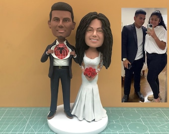 May include: A custom wedding cake topper featuring a bride and groom. The groom is depicted as a superhero, revealing a Spider-Man design on his shirt. The bride wears a white wedding dress and holds a bouquet of red roses.