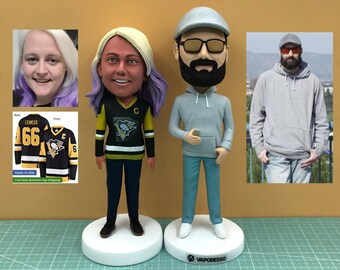 May include: Custom bobblehead figurines of a couple, each standing on a white base. The woman's figurine wears a black and gold hockey jersey. The man's figurine wears a gray hoodie and cap. Additional images of the couple are included.