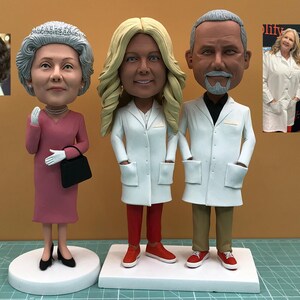May include: Three bobblehead figurines: a woman in a pink dress, a woman in a white lab coat and red pants, and a man in a white lab coat and khaki pants. Each figure has a large head and is standing on a white base. A small photo is in the background.