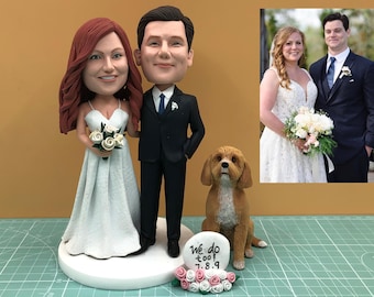 May include: A wedding cake topper featuring a bride and groom figurine, a dog figurine, and a small sign that reads "We do too! 7.8.9". The bride wears a white dress and holds a bouquet. The groom wears a black suit.