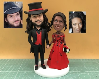 May include: A custom wedding cake topper featuring a couple holding hands. The groom wears a black suit with a red vest and a top hat. The bride wears a red gown with black floral accents and holds a bouquet.