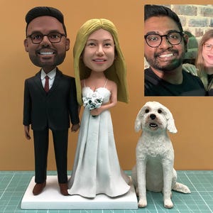 May include: A custom wedding cake topper featuring a bride and groom figurine, a white dog, and a photo of the couple. The groom wears a black suit and the bride a white dress, holding a bouquet. The dog is white.