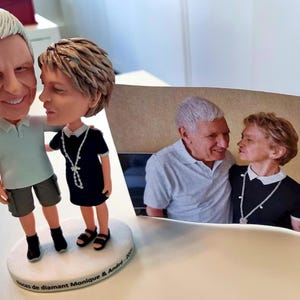 Könnte beinhalten: Eine individuelle Wackelkopf-Figur eines Paares, mit einem Foto des Paares im Hintergrund. Die Figur zeigt einen Mann in einem hellblauen Hemd und Shorts und eine Frau in einem schwarzen Kleid und Sandalen. Die Basis der Figur hat Text.