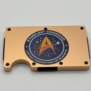 Könnte beinhalten: Eine goldfarbene Metallgeldbörse mit dem Starfleet Command-Logo. Das Logo ist ein kreisförmiges Emblem mit einem Stern, einem Planeten und dem Text "Starfleet Command United Federation of Planets."