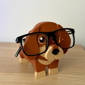 Może przedstawiać: Brązowy drewniany uchwyt na okulary w kształcie psa z beżowym pyskiem i łapami. Pies nosi okulary w czarnych oprawkach. Uchwyt stoi na drewnianej powierzchni.