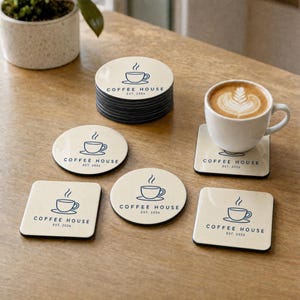 May include: A collection of round and square coasters in cream with a dark blue graphic of a coffee cup and the text COFFEE HOUSE EST. 2026. One square coaster supports a white ceramic mug filled with coffee featuring latte art on the surface.