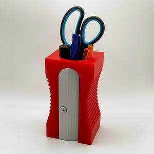 May include: Red plastic pencil holder with a built-in pencil sharpener. The holder has a ribbed design and is filled with pens, pencils, and a pair of scissors.