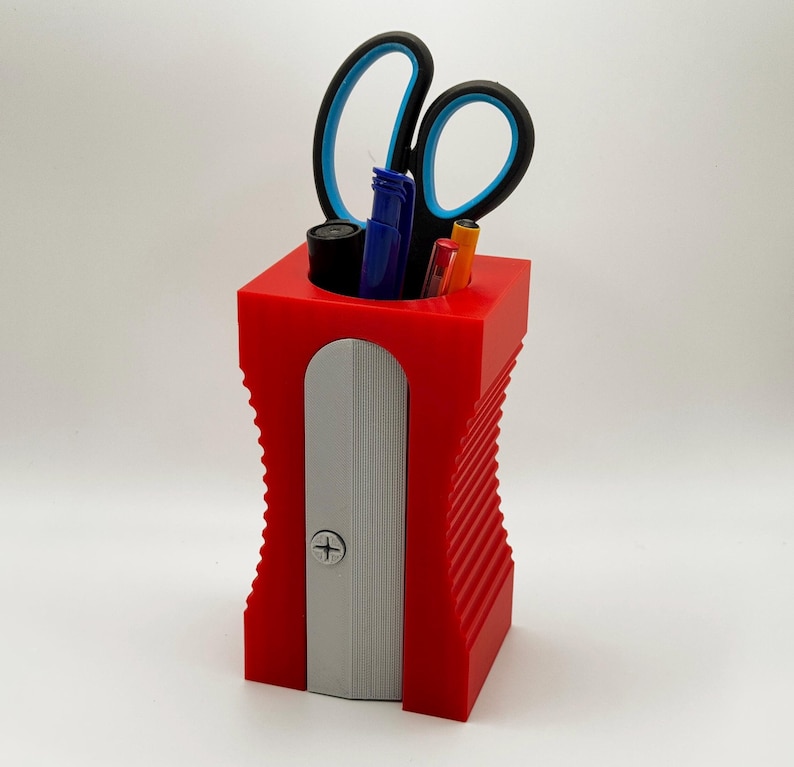 May include: Red plastic pencil holder with a built-in pencil sharpener. The holder has a ribbed design and is filled with pens, pencils, and a pair of scissors.
