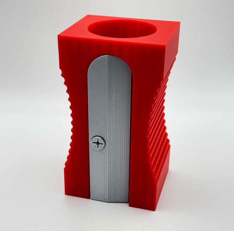 May include: A red plastic pencil sharpener with a silver metal blade. The sharpener has a cylindrical opening for inserting pencils and a ribbed design on the sides.